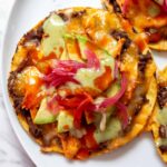 a bright and colorful chicken tostada with green salsa and pink pickled onions on a white plate.
