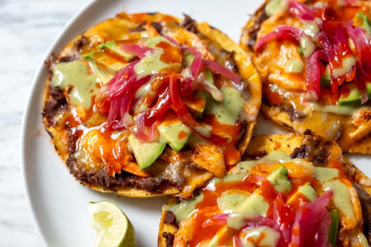 a white plate filled with 3 cheesy chicken tostadas topped with avocado, pink onions, red salsa and a wedge of lime.