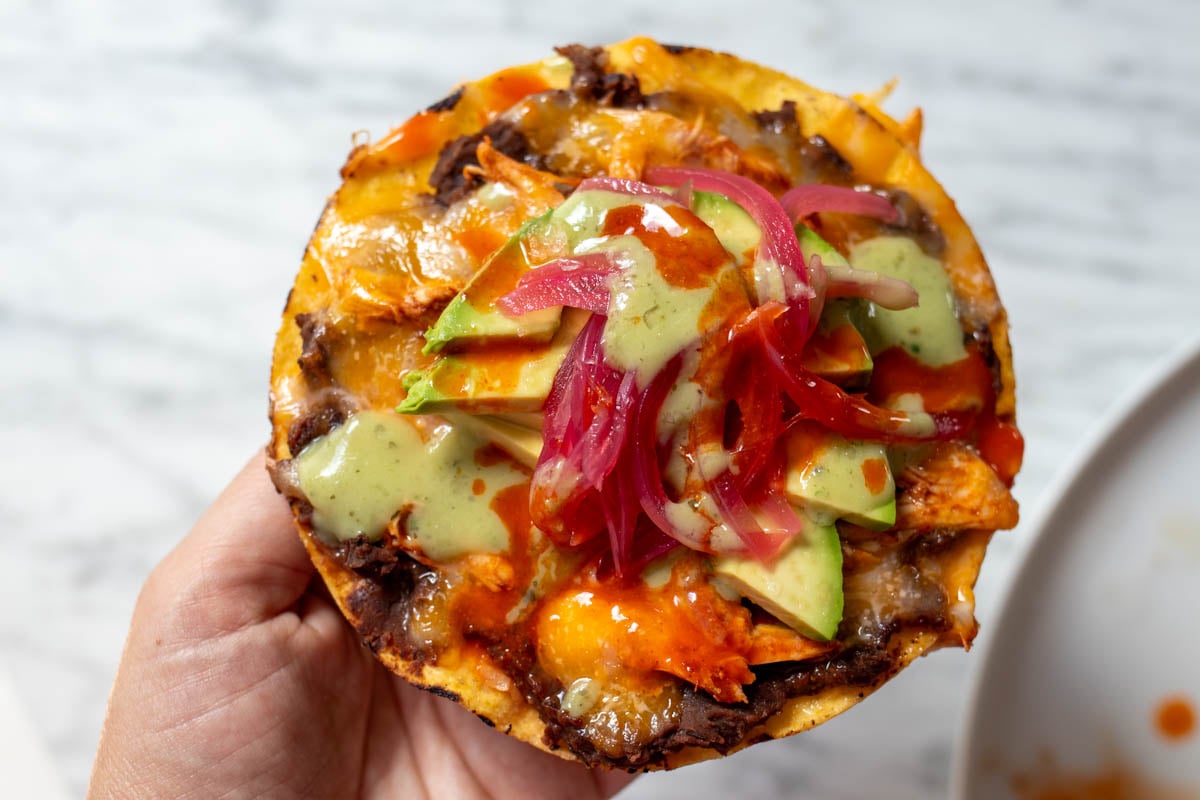 a hand holding a singular chicken tostada with melted cheese, avocado, and pink pickled onions.