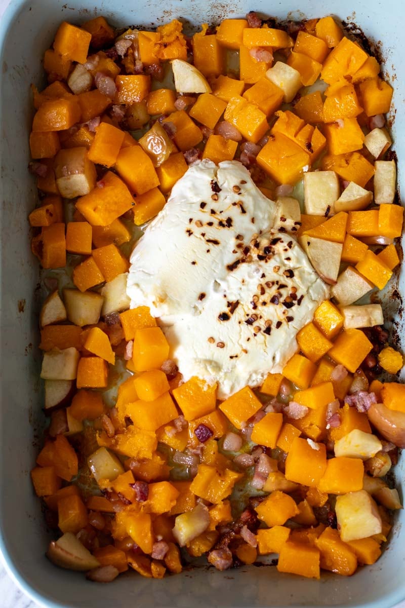 a blue baking dish full of cubed butternut squash, pancetta, apples and two logs of goat cheese topped with seasoning.