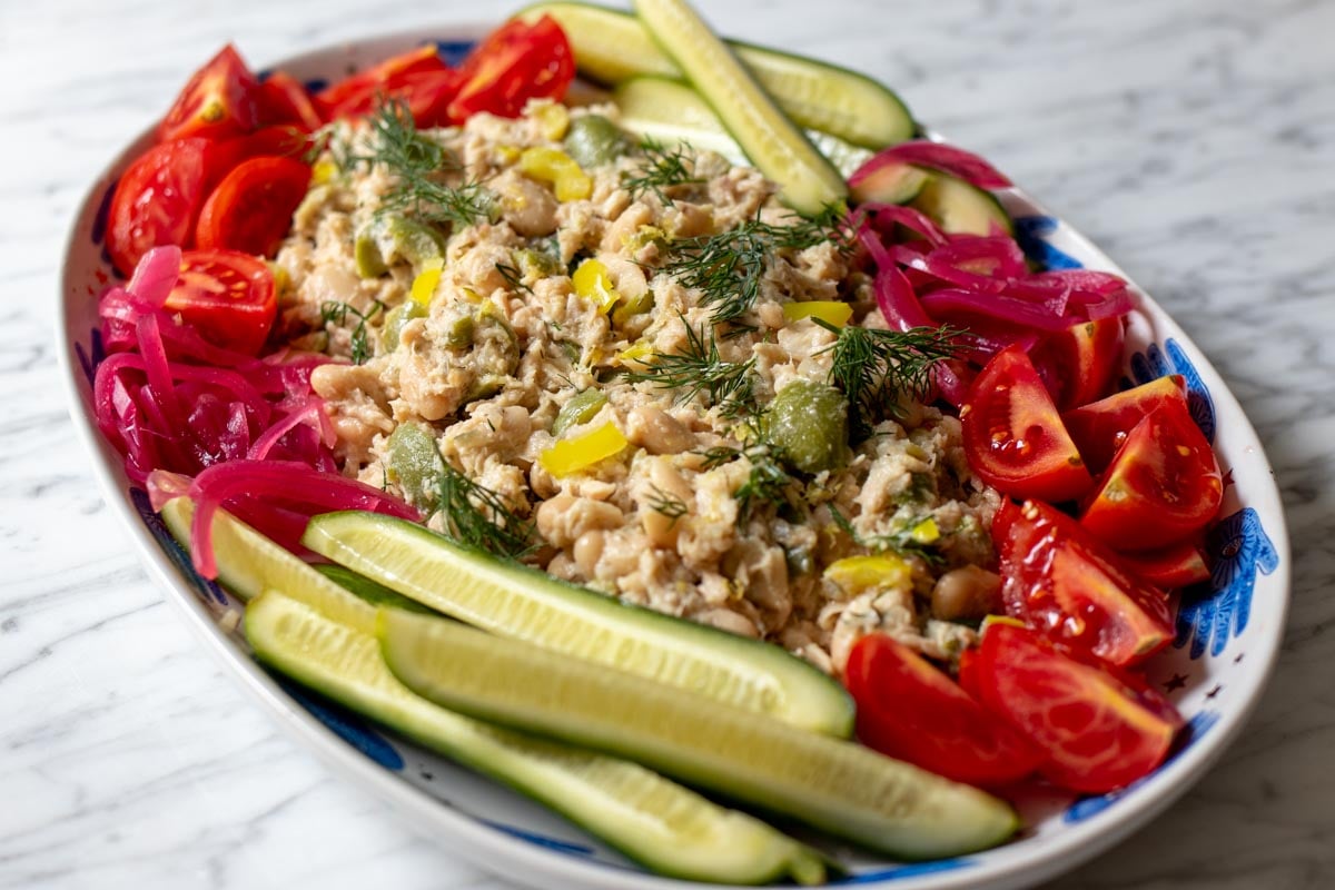 a white platter with greek inspired by hands lining the border filled with lemony tuna salad and colorful chopped veggies surrounding it.