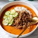 a broth bowl full of crispy meat, bright red curry broth, white rice and green cucumbers.