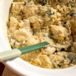 a spoon scooping up some creamy chicken orzo out of a cream colored crockpot.
