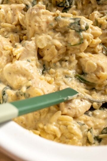 a spoon scooping up some creamy chicken orzo out of a cream colored crockpot.