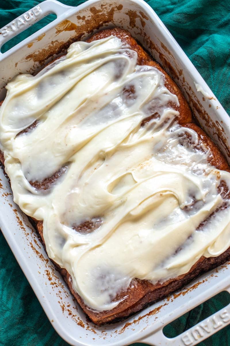 a baking dish full of cinnamon rolls with cream cheese icing on top of a wooden table.