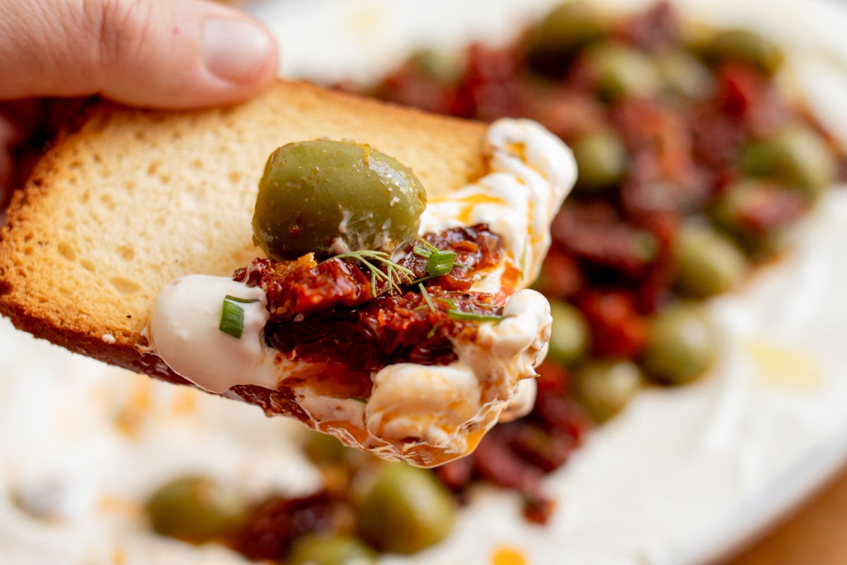 a hand scooping up some yogurt feta dip with an olive and sun dried tomatoes on top.